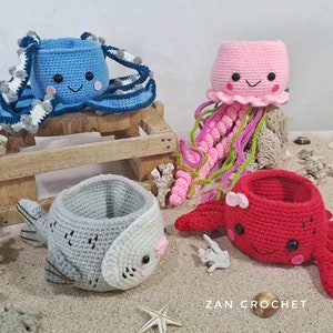 Puede incluir: Cuatro contenedores de animales de peluche tejidos a crochet en forma de pulpo azul, medusa rosa, pez gris y cangrejo rojo. Los contenedores están sentados en una caja de madera y una superficie arenosa. El texto "ZAN CROCHET" es visible en la esquina inferior derecha de la imagen.