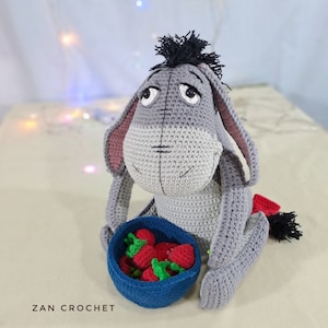 May include: A gray crocheted stuffed donkey with black yarn for its mane and tail, sitting on a white surface. The donkey is holding a blue bowl filled with red crocheted strawberries.
