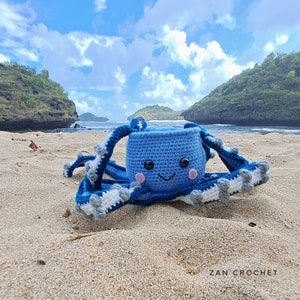 Puede incluir: Un pulpo de crochet azul con una cara sonriente y detalles blancos y grises en sus tentáculos. El pulpo está sentado en una playa de arena con un océano azul y colinas verdes en el fondo.