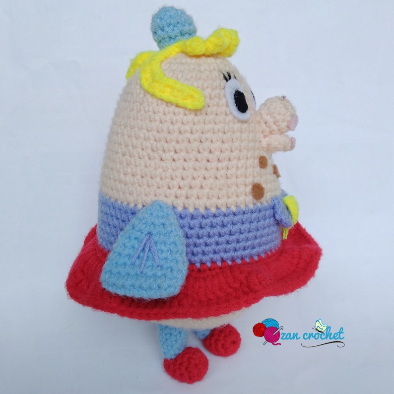 Peut inclure: Un jouet en peluche crochet&eacute; en forme de personne avec un corps rose, une jupe bleu et violet, des chaussures rouges et des cheveux jaunes. Le jouet porte un petit chapeau bleu sur la t&ecirc;te et un nez brun.