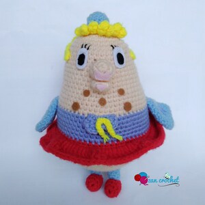 Peut inclure: Un jouet en peluche crochet&eacute; en forme de personne avec une couronne jaune, une robe bleue et rouge et des taches brunes. Le jouet a un nez rose et de grands yeux.