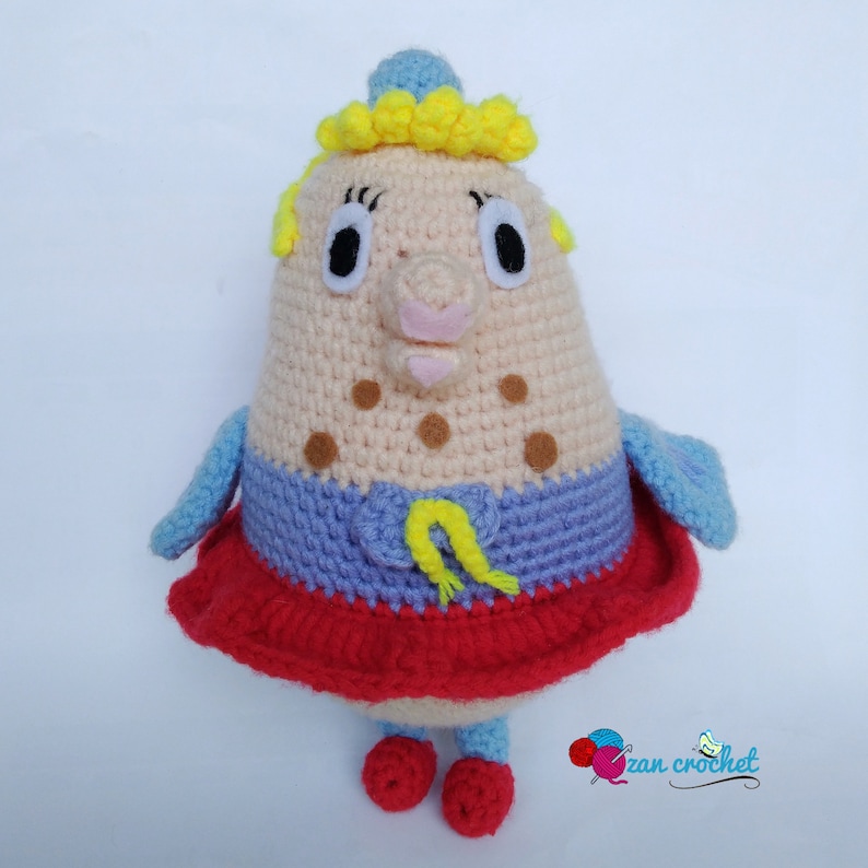 Peut inclure: Un jouet en peluche crochet&eacute; en forme de personne avec une couronne jaune, une robe bleue et rouge et des taches brunes. Le jouet a un nez rose et de grands yeux.