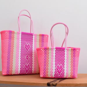 May include: Two woven tote bags in varying sizes, featuring a vibrant striped pattern in shades of hot pink, light pink, white, and soft orange. Each bag has two matching woven handles and a subtle heart-shaped design integrated into the weave.
