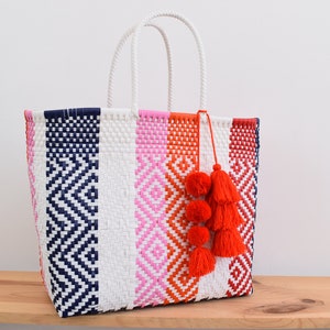 May include: A white woven tote bag with blue, pink, and orange stripes and geometric patterns. The bag has three red pom-poms attached to the handle.
