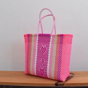 May include: A rectangular tote bag constructed from woven plastic strips in a vibrant colour palette of hot pink, magenta, white, and light beige. The bag features two matching pink woven handles and a repeating vertical striped pattern throughout.