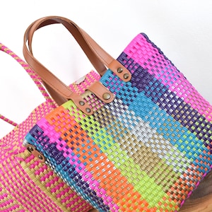 May include: Two woven tote bags in vibrant colors. One is primarily pink with a matching handle, the other is a rainbow of colors including pink, orange, yellow, green, blue, and purple. Both bags have brown leather handles.