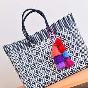 May include: A woven tote bag with a geometric pattern in navy blue and white. The bag has navy blue handles and is adorned with a colorful tassel of red, orange, blue, purple, and pink pom-poms. The bag is sitting on a wooden surface.