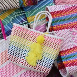 May include: A selection of brightly coloured woven tote bags. The bags display a rainbow of hues, including pink, yellow, blue, and white. One bag has a yellow tassel. The handles are white and pink.