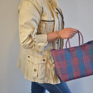 May include: A woven tote bag in shades of blue and red is held. The bag has two braided handles and a rectangular shape. The person is wearing a tan jacket and blue jeans. The bag is suitable for shopping.