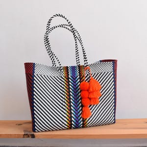 May include: Woven tote bag with black and white chevron pattern, accented with red, blue, and yellow stripes. Features black and white handles and a cluster of orange tassels. Sits on a wooden surface.