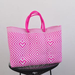 May include: A rectangular tote bag featuring a woven pattern of hot pink and white geometric diamond shapes. The bag has two matching hot pink braided handles attached to the top edge, providing a structured shape and a vibrant colour contrast.