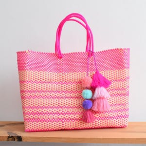 May include: A woven tote bag in shades of pink and peach, featuring a rectangular shape and sturdy handles. The bag is adorned with a decorative tassel of pink, purple, and blue pom-poms. The bag is sitting on a wooden surface.