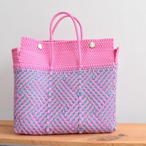 May include: A pink and blue woven tote bag with a rectangular shape. The top section is solid pink, while the lower section features a chevron pattern in blue, green, and purple. The bag has two handles and two silver snap closures.