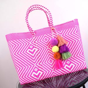 May include: A vibrant pink and white woven tote bag with a geometric pattern. The bag features matching handles and is adorned with a colorful tassel of pom-poms in shades of pink, purple, yellow, orange, and green. The bag is sitting on a glass table.