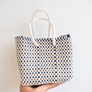 May include: A woven tote bag with a checkered pattern in navy blue, tan, and white. The bag has white rope handles and is held up against a white background. The bag is a small size.