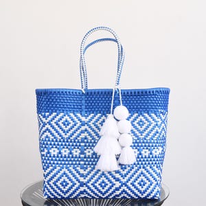 May include: A woven tote bag featuring a blue and white geometric diamond pattern. The bag has two matching woven handles and is decorated with a cluster of three white pom-poms and three white tassels hanging from one of the handles.