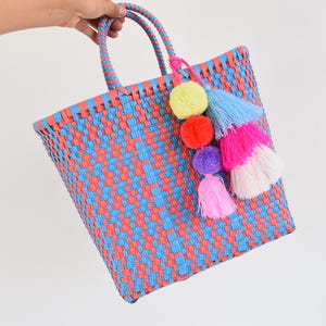 May include: A woven tote bag in a blue and coral pattern, held by a hand. The bag has two handles and a colorful tassel with yellow, blue, red, purple, pink, and white accents. The bag is made of a woven material.