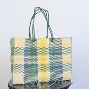 May include: A rectangular tote bag constructed from woven plastic strips in a plaid pattern of cream, sage green, light blue, and bright yellow. The bag features two matching green braided handles attached to the top edge of the structured body.