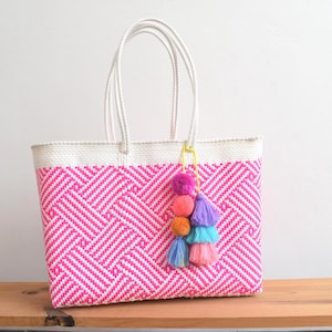 May include: A woven tote bag with a pink and white geometric pattern. The bag has white handles and a white top border. A colorful tassel with pink, orange, purple, and blue accents hangs from the handle. The bag is on a wooden surface.