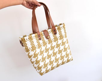 Puede incluir: Un bolso tote tejido con un estampado de pata de gallo en blanco y dorado. El bolso tiene asas de cuero marrón y una correa de cierre a juego. El bolso está sostenido por una mano sobre un fondo blanco.