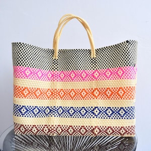 May include: Woven tote bag with a checkered black and white top section, and horizontal stripes of pink, orange, blue, and brown. The bag has beige handles and is made of a natural fiber material.