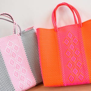 May include: Three woven tote bags in bright colours. One bag is pink and grey, another is pink and orange, and the third is orange and pink. The bags have woven handles and a diamond pattern on the front.