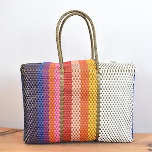May include: A rectangular woven tote bag with a vibrant striped design. The bag's pattern includes blue, purple, pink, orange, and yellow stripes. It has olive green handles and a white and blue woven side.