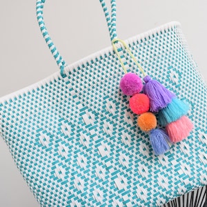 May include: A woven tote bag with a turquoise and white diamond pattern. The bag has turquoise handles and a colourful tassel with pink, purple, orange, and blue pom-poms and tassels. A stylish accessory.