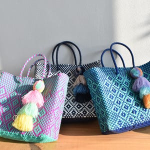 May include: Three woven tote bags with geometric patterns and matching handles. Each bag features a colourful tassel. The bags are in shades of blue, pink, and black, displayed on a wooden surface against a white wall.