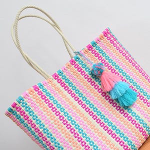 May include: A woven tote bag featuring a multi-coloured striped pattern in shades of bright pink, turquoise, peach, and cream. The bag includes two braided cream-coloured handles and a decorative multi-layered tassel in pink and turquoise.