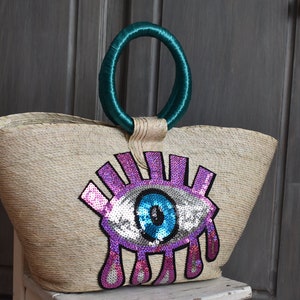 Puede incluir: Un bolso de paja con un gran diseño de ojo colorido hecho de lentejuelas. El ojo es azul, rosa y plateado. El bolso tiene un asa de tela turquesa.