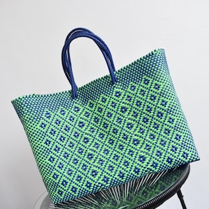 May include: A woven tote bag in a geometric pattern of green and navy blue diamonds. The bag has navy blue handles and is sitting on a small, round table. The bag is made of a durable, woven material.