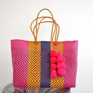 May include: A vibrant, woven tote bag with a geometric pattern in pink, yellow, and blue. The bag features twisted rope handles and a decorative tassel with three pink pom-poms. This colorful bag is perfect for carrying groceries or beach essentials.
