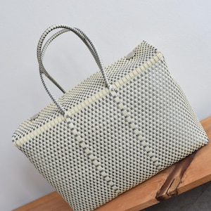 May include: A grey and cream woven tote bag with a checkered pattern. The bag features two grey handles and a rectangular design. The bag is made from a durable, woven material, suitable for carrying items such as groceries.