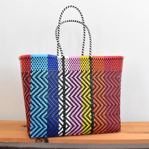 May include: A rectangular tote bag constructed from woven plastic strips in a vibrant chevron pattern. The bag features vertical stripes of blue, black, white, pink, yellow, orange, and red, complemented by two black and white twisted rope handles.