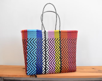 May include: A rectangular tote bag constructed from woven plastic strips in a vibrant chevron pattern. The bag features vertical stripes of blue, black, white, pink, yellow, orange, and red, complemented by two black and white twisted rope handles.