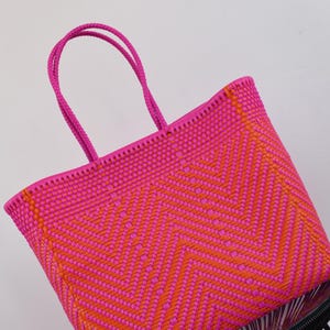 May include: A woven tote bag featuring a vibrant hot pink and bright orange chevron pattern. The bag is constructed from thin plastic strips woven into a tight, structured design with two matching hot pink braided handles at the top.