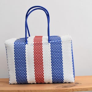 May include: A rectangular woven tote bag with blue, white, and red vertical stripes. The bag has blue rope handles. The bag is a vibrant accessory. It is sitting on a wooden surface.