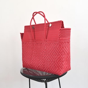 May include: A bright red tote bag constructed from woven plastic strips featuring a diamond pattern on the lower section and a solid weave on the upper portion. The bag includes two thin red handles and two silver-toned circular metal fasteners.