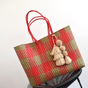 May include: A woven tote bag with red and olive green plaid pattern. The bag has red handles and is adorned with a beige tassel and pom-pom embellishment. The bag is sitting on a black chair.