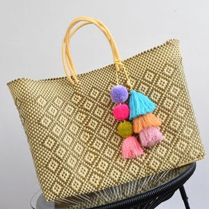 May include: Woven tote bag with a diamond pattern in shades of beige and brown. The bag has yellow handles and is adorned with a colorful tassel featuring purple, pink, blue, orange, and pink pom-poms. A stylish and practical accessory.