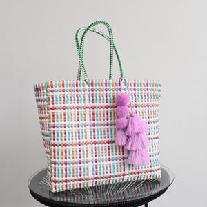 May include: A rectangular woven tote bag featuring a multi-coloured grid pattern in white, orange, blue, and purple. The bag has two green woven handles and is decorated with a hanging accessory consisting of three purple pom-poms and a tassel.