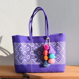 May include: A woven purple tote bag with white diamond pattern. The bag has matching purple handles and a decorative tassel with pink, orange, teal, and purple pom-poms. The bag is sitting on a wooden surface.