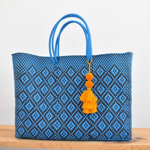 May include: Blue and black woven tote bag with diamond pattern. The bag has blue handles and a decorative orange tassel with two pom-poms. The bag is sitting on a wooden surface.