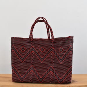 May include: A woven tote bag in a dark burgundy color with a geometric pattern in black and red. The bag has two handles made of twisted cord. The bag is sitting on a wooden surface.