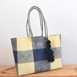 May include: Woven tote bag with a checkered pattern in cream and navy blue. The bag has matching handles and a navy tassel. The bag is sitting on a wooden surface.