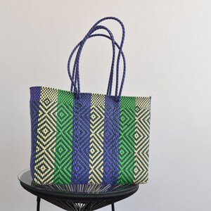 May include: A woven tote bag with a geometric pattern in shades of green, purple, and white. The bag has two purple handles.