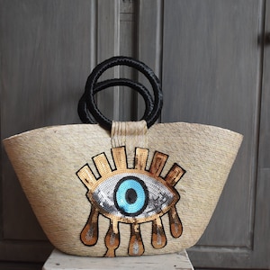 Puede incluir: Un bolso de paja con un gran diseño de ojo azul hecho de lentejuelas. El ojo tiene detalles dorados y una pupila negra. El bolso tiene asas negras.