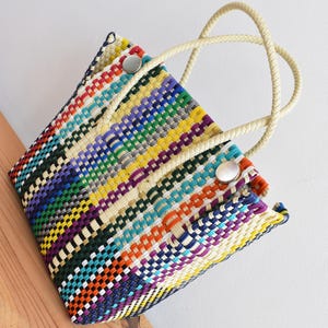 Può includere: Una borsa tote intrecciata multicolore di forma rettangolare. La borsa presenta un vivace mix di colori, tra cui rosso, giallo, blu e verde, disposti a strisce orizzontali. Ha manici in corda color crema e chiusure con bottoni argentati.