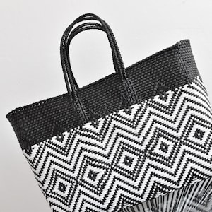 May include: Black and white woven tote bag with a geometric pattern. The bag has a rectangular shape with a black top and handles. The lower portion features a repeating white and black zigzag design.